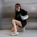 Person taking a mirror selfie wearing a black outfit with a white bag, sitting on a gray floor.