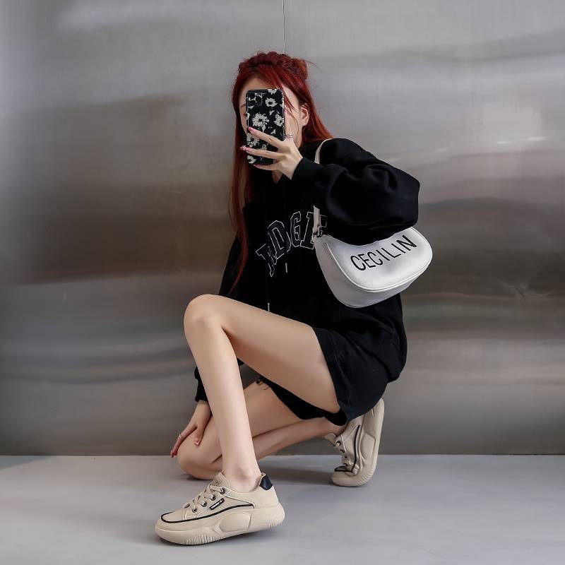 Person taking a mirror selfie wearing a black outfit with a white bag, sitting on a gray floor.