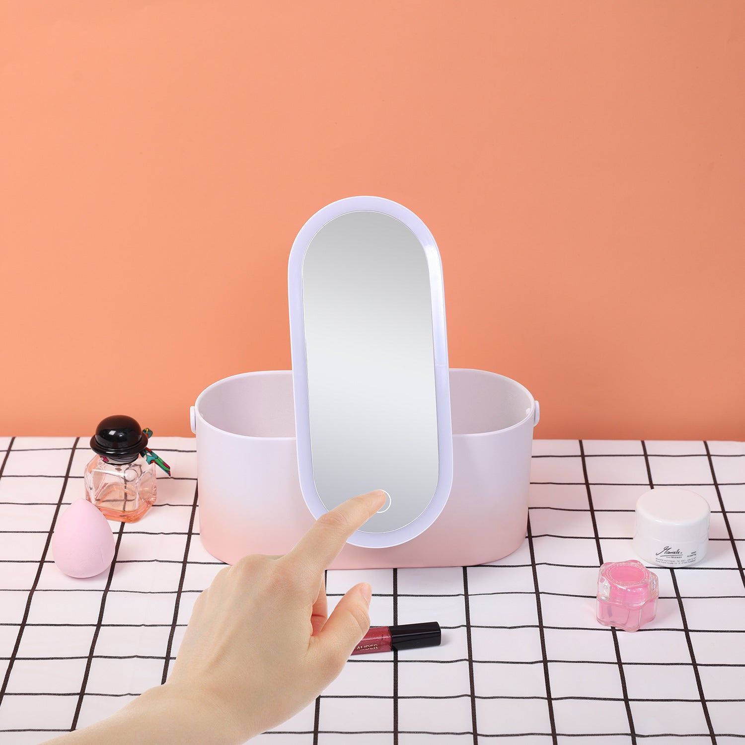 Hand holding a small round mirror over a pink container with cosmetic items on a checkered tablecloth.