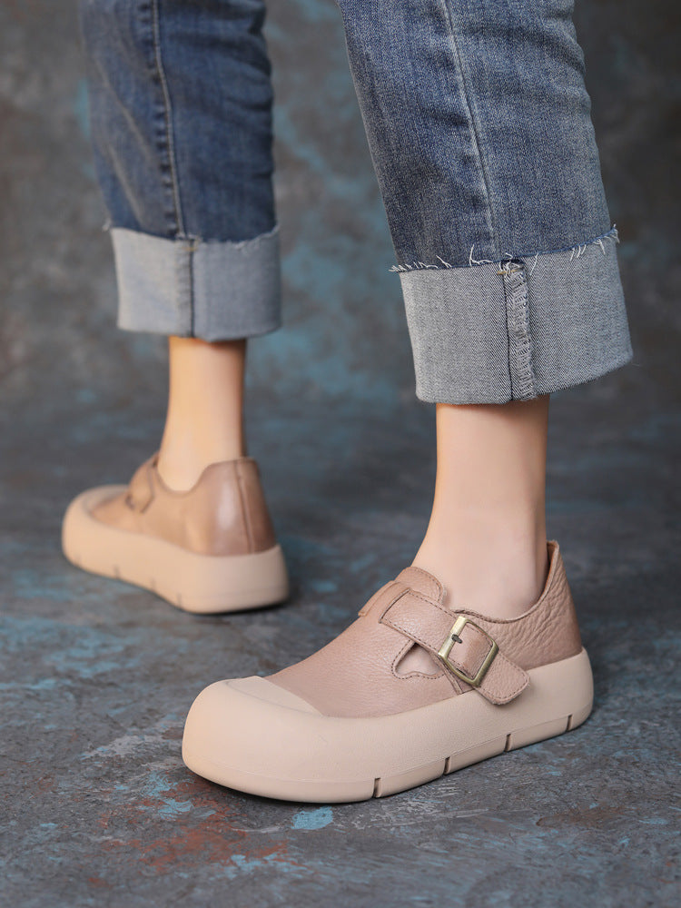 Beige shoes with a buckle worn with rolled-up jeans on a textured gray floor.