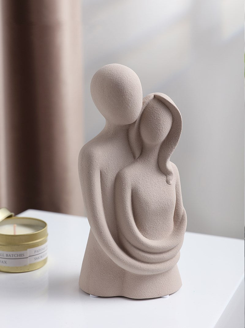 Ceramic sculpture of two figures embracing on a white surface with a candle in the background.
