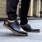 Black leather shoes with brown soles worn by a person on a pavement.