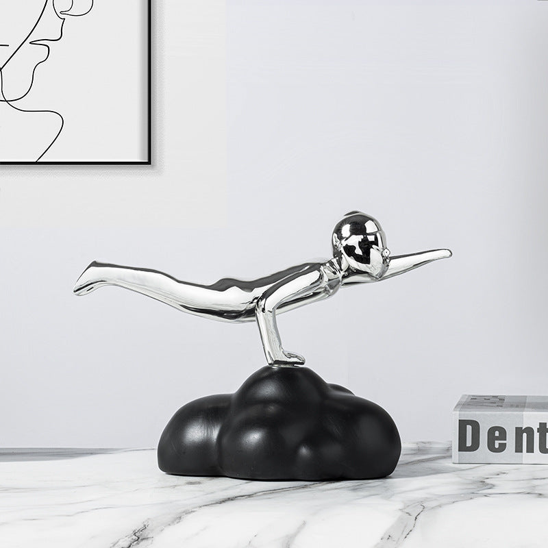 Decorative sculpture of a silver diving figure on a black cloud base, with a white marble surface and abstract art in the background.