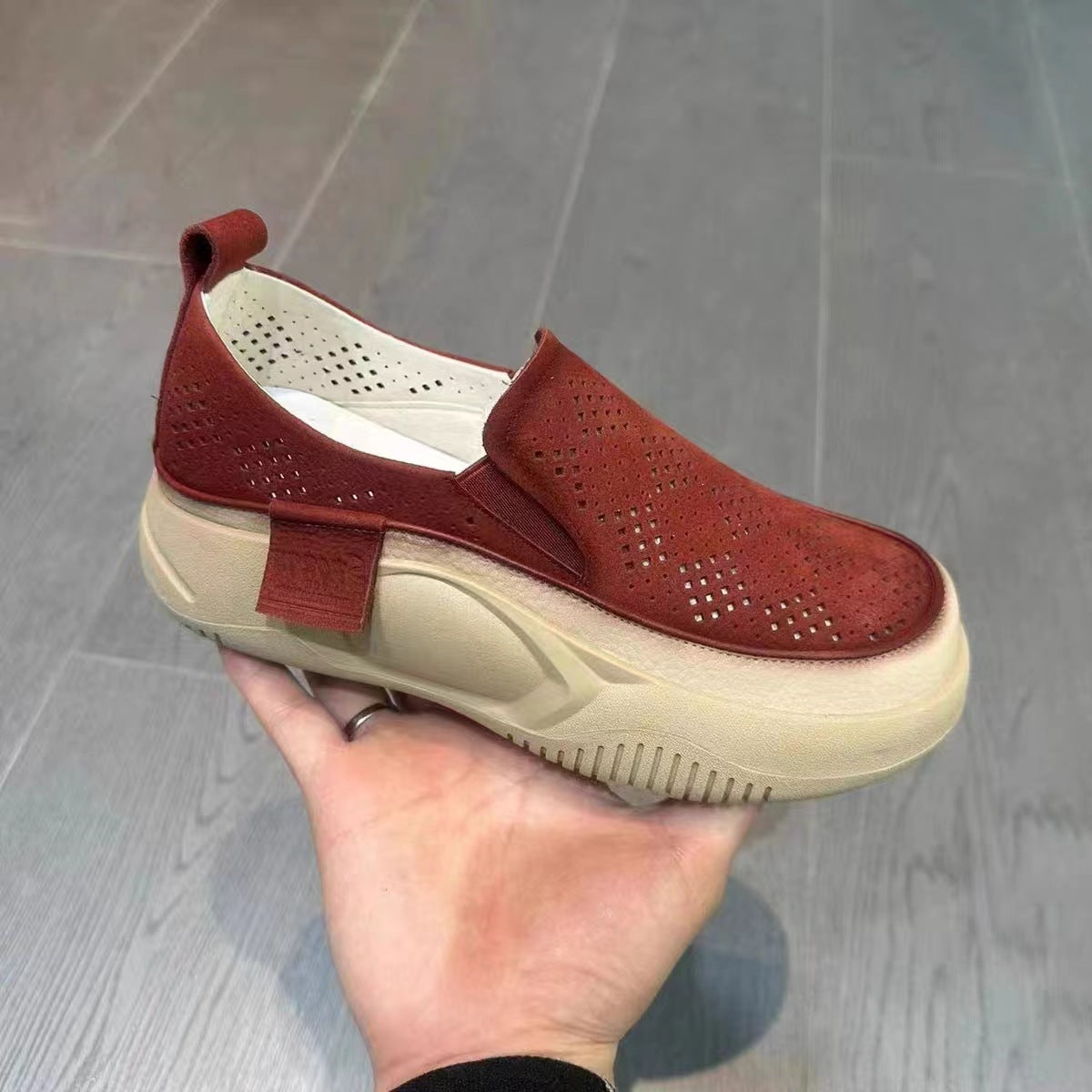 Red slip-on shoe held in a hand against a gray floor background