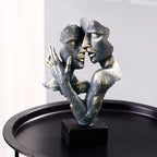 Bronze sculpture of two faces embracing on a black base against a gray background