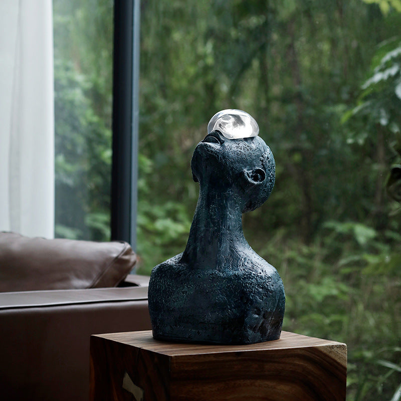 Decorative sculpture with a spherical object on top, placed on a wooden surface with a blurred natural background.