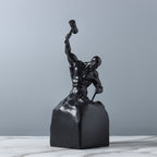 Black sculpture of a muscular figure on a marble surface with a gray background