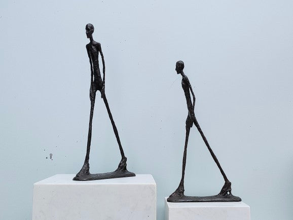Two bronze sculptures of humanoid figures on white plinths against a light blue background