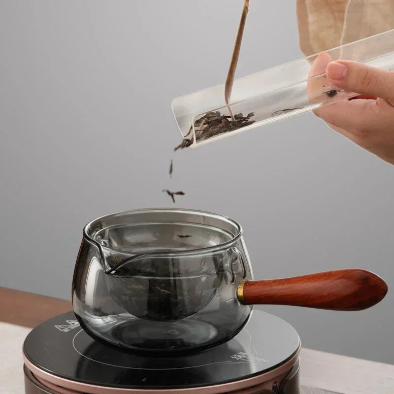 Person pouring tea leaves into a glass teapot on a gray background