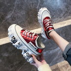 Red sneakers with white soles held by a person against a dark stone wall.